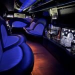 Interior of a 22-passenger SUV limo featuring curved leather seating, crystal decanters, and a glowing bar setup under ambient blue lighting.