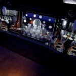 Close-up view of a luxury bar inside an SUV limo, showcasing crystal decanters, glass holders, and reflective mirrored back panels with ambient lighting.